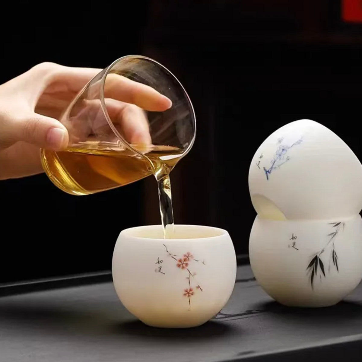 Person pouring tea from a glass teapot into two white ceramic cups with artistic designs on a dark surface.