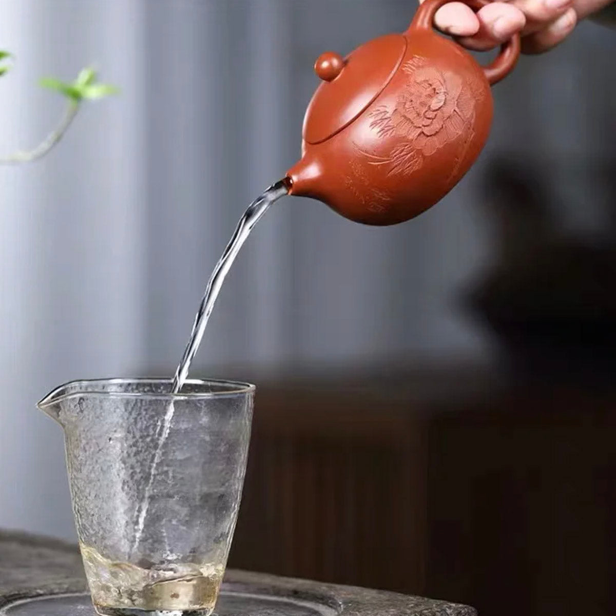 Red ceramic teapot pouring water into a clear glass with a blurred background