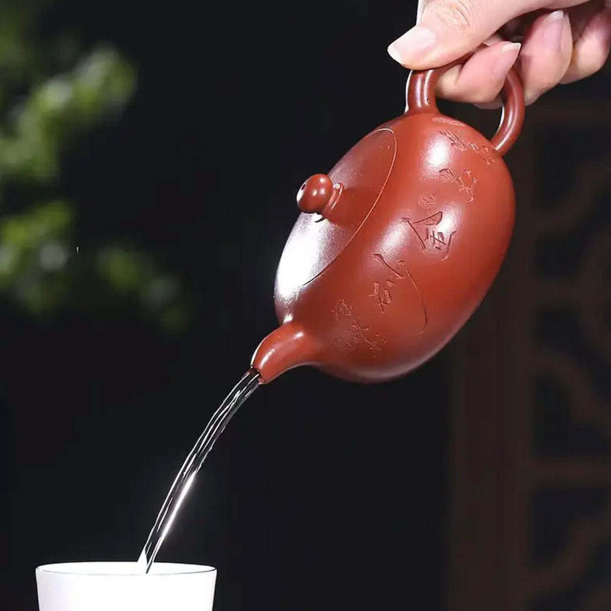 Red ceramic teapot being used to pour tea into a white cup against a dark background