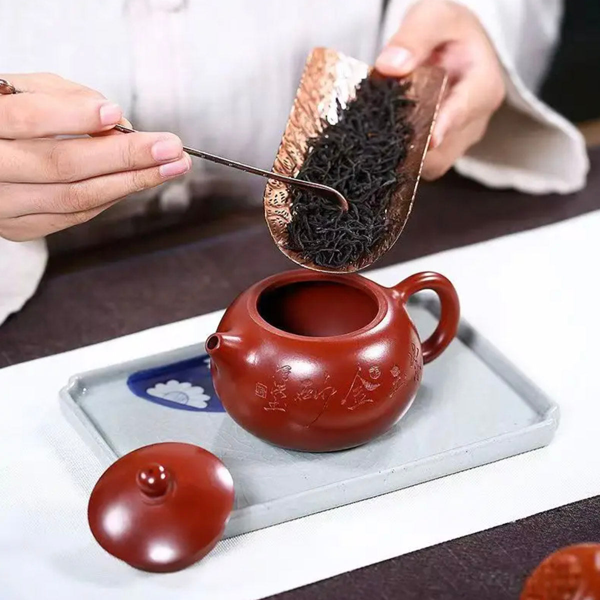 Person adding tea leaves to a red teapot on a white tray