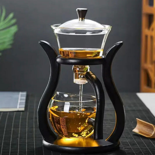 Glass teapot with black wooden handle and stand on a dark surface.