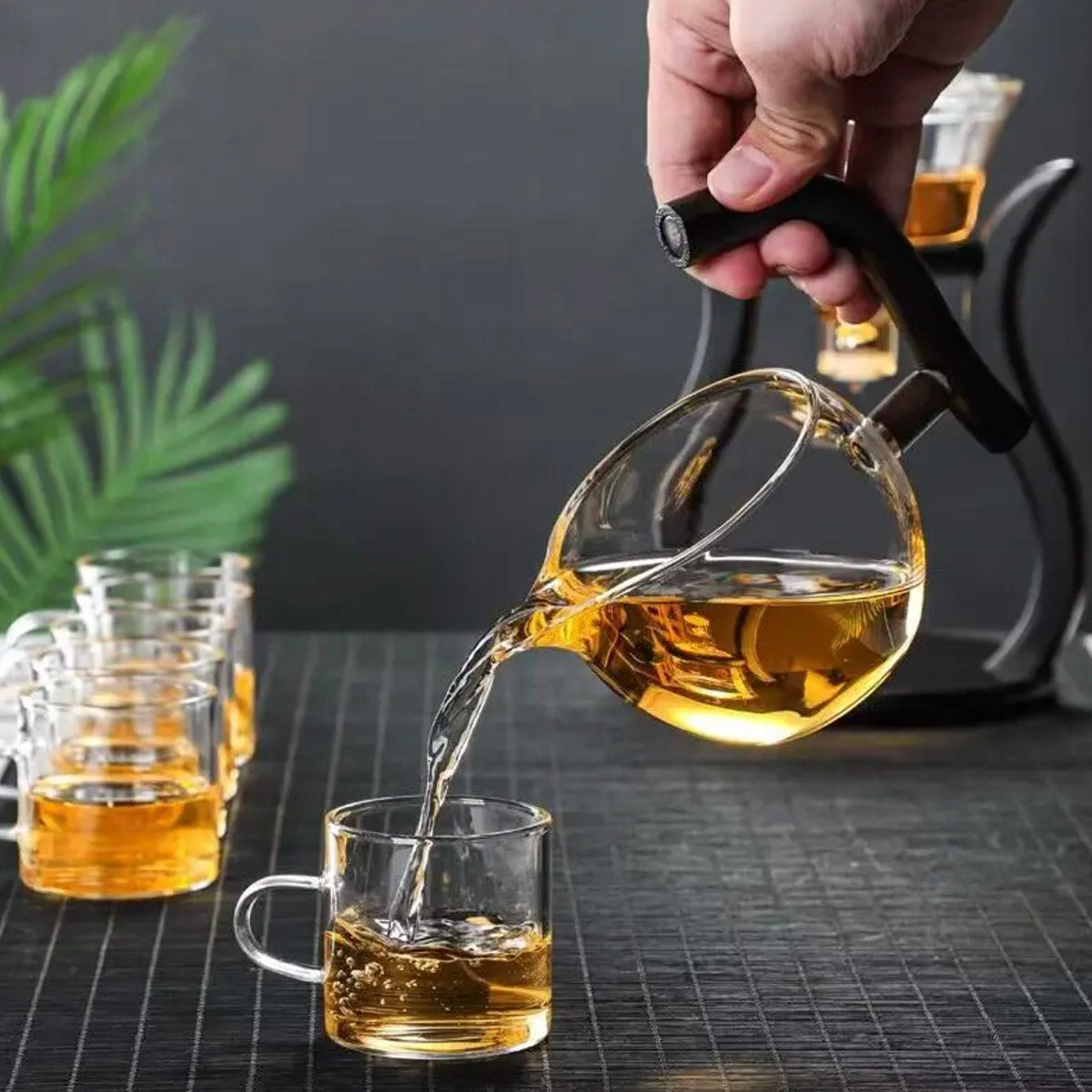 Person pouring tea from a glass teapot into a clear mug on a dark surface with a plant in the background.